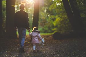 Vater und Tochter halten sich im Gegenlicht an den Händen und gehen gemeinsam einen herbstlichen Waldweg entlang